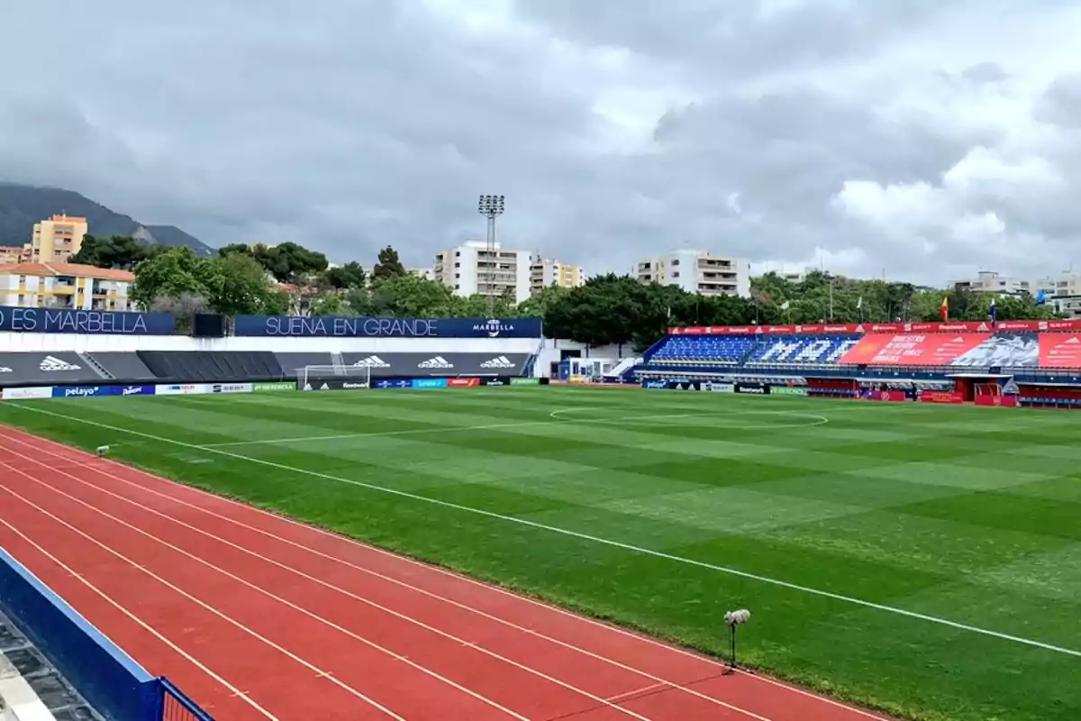 Estadio de fútbol con pista de atletismo alrededor, gradas azules y rojas, césped bien cuidado y edificios residenciales al fondo bajo un cielo nublado Estadio de fútbol con pista de atletismo alrededor, gradas azules y rojas, césped bien cuidado y edificios residenciales al fondo bajo un cielo nublado
