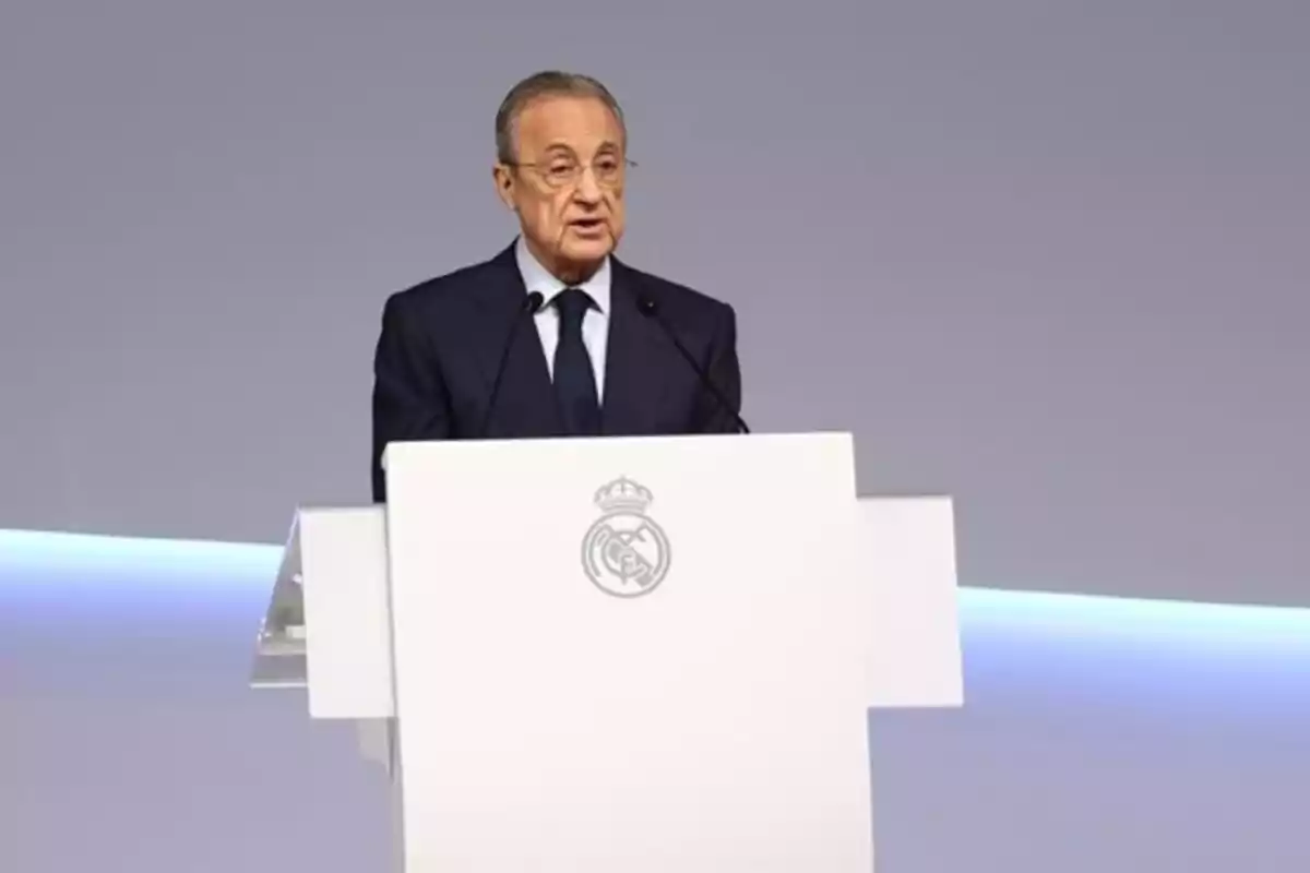 Hombre de traje hablando en un podio blanco con el escudo del Real Madrid sobre un fondo gris