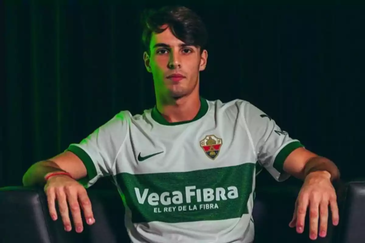 Joven con camiseta blanca y verde del Elche CF sentado en un sillón negro con fondo oscuro Joven con camiseta blanca y verde del Elche CF sentado en un sillón negro con fondo oscuro