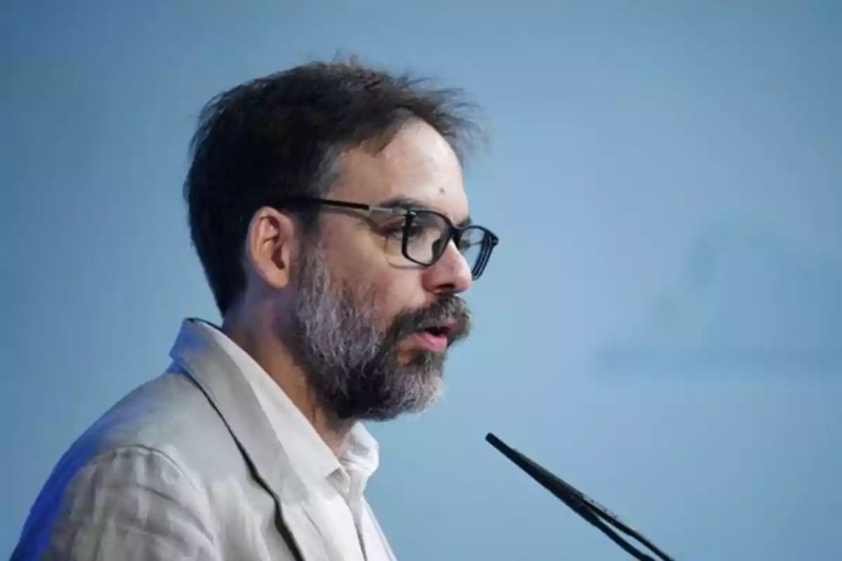 Hombre de barba y gafas hablando frente a un micrófono con fondo azul Hombre de barba y gafas hablando frente a un micrófono con fondo azul