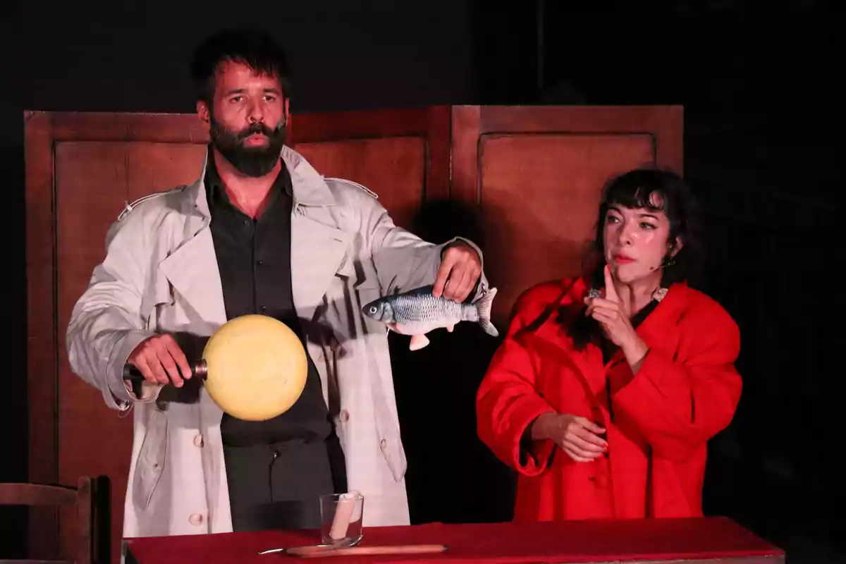 Un hombre con barba y gabardina sostiene un objeto redondo amarillo y un pez de juguete mientras una mujer con abrigo rojo lo observa y hace un gesto de silencio