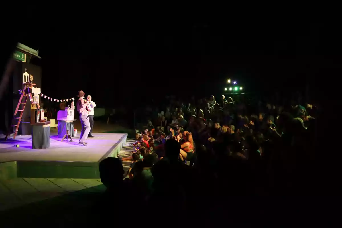 Dos artistas realizan un espectáculo en un escenario iluminado mientras una gran audiencia observa y aplaude desde las gradas en un ambiente nocturno Dos artistas realizan un espectáculo en un escenario iluminado mientras una gran audiencia observa y aplaude desde las gradas en un ambiente nocturno