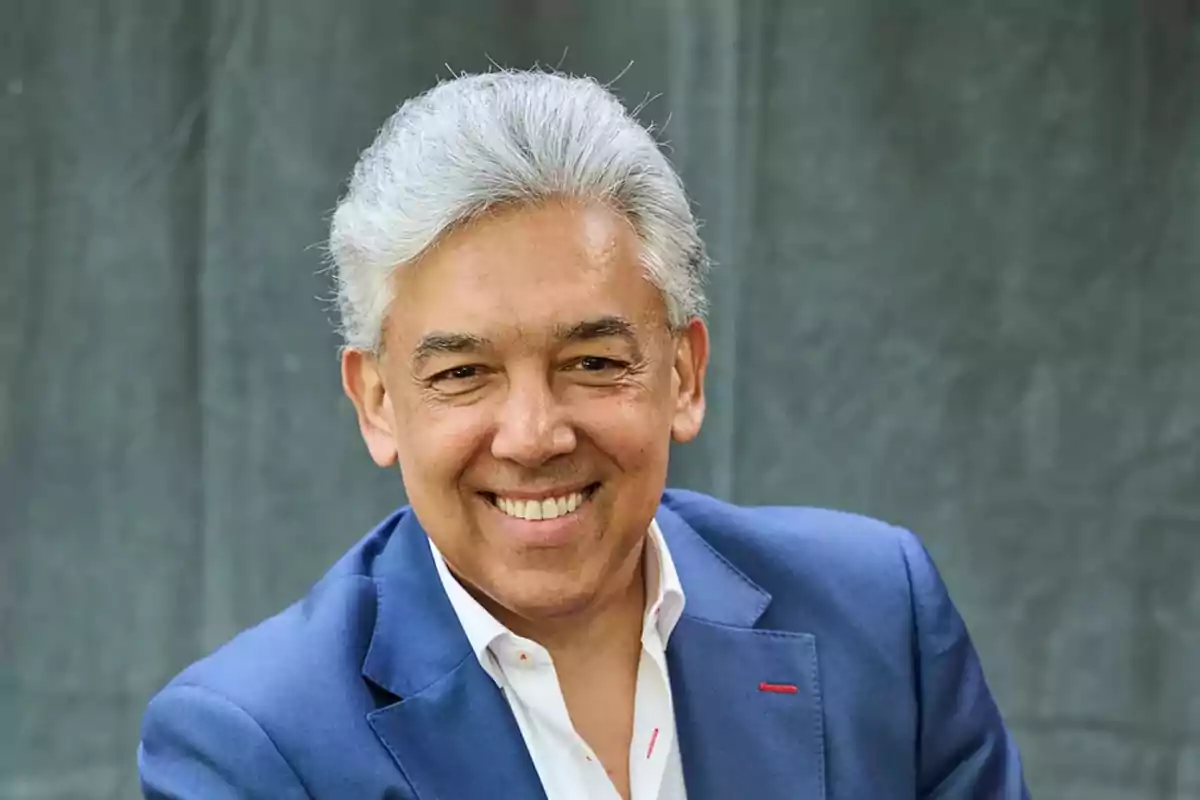 Hombre de cabello canoso y sonrisa amplia vistiendo saco azul frente a un fondo gris