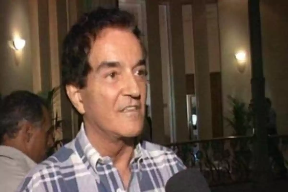 Hombre de cabello oscuro con camisa de cuadros hablando frente a un micrófono en un lugar interior iluminado Hombre de cabello oscuro con camisa de cuadros hablando frente a un micrófono en un lugar interior iluminado