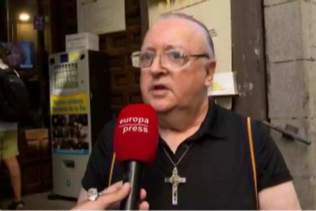Persona de cabello corto y gafas siendo entrevistada en la calle con un micrófono rojo de europa press lleva una cruz colgada al cuello y viste camiseta negra Persona de cabello corto y gafas siendo entrevistada en la calle con un micrófono rojo de europa press lleva una cruz colgada al cuello y viste camiseta negra