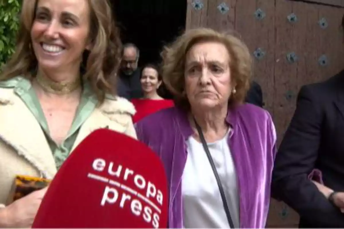 Dos mujeres están frente a un micrófono de Europa Press, una sonríe y la otra tiene expresión seria, al fondo hay más personas y una puerta de madera Dos mujeres están frente a un micrófono de Europa Press, una sonríe y la otra tiene expresión seria, al fondo hay más personas y una puerta de madera