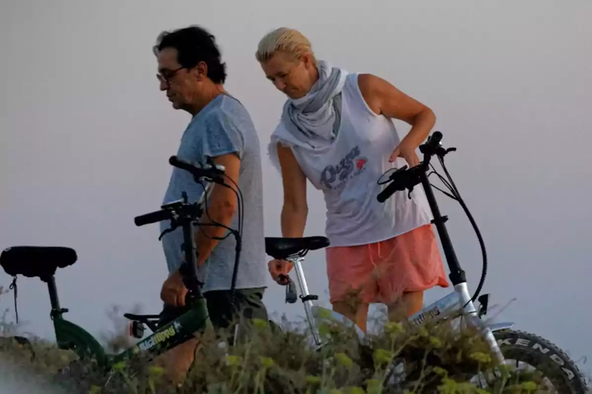 Dos personas caminan junto a sus bicicletas en un entorno natural con vegetación baja y cielo despejado Dos personas caminan junto a sus bicicletas en un entorno natural con vegetación baja y cielo despejado
