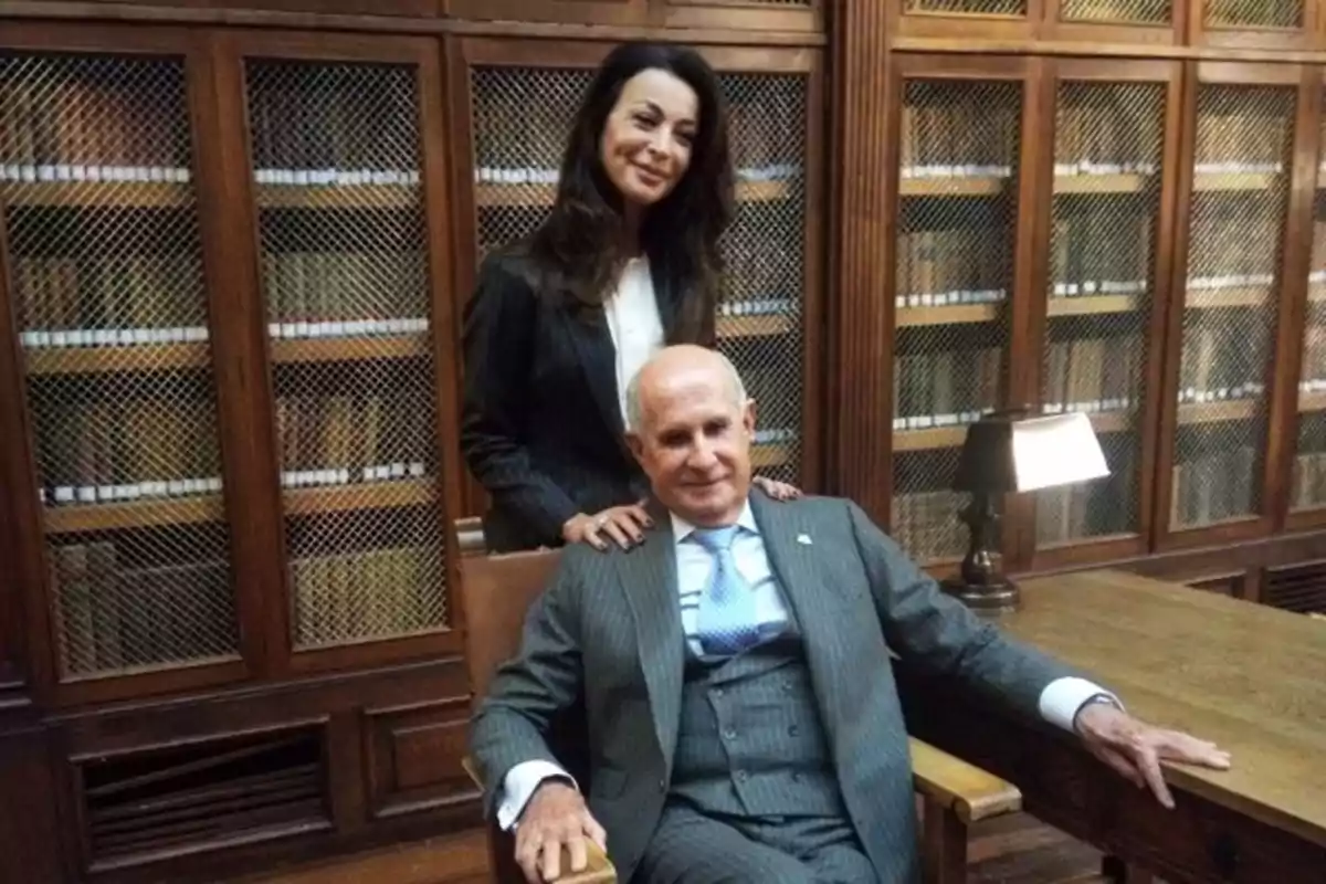 Una mujer de pie y un hombre sentado posan juntos en una biblioteca antigua con estanterías de madera llenas de libros al fondo Una mujer de pie y un hombre sentado posan juntos en una biblioteca antigua con estanterías de madera llenas de libros al fondo