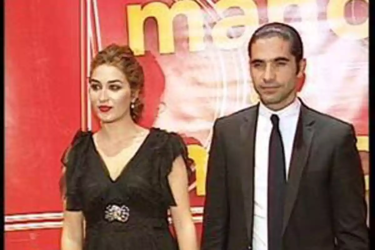 Una mujer con vestido negro y un hombre con traje posan juntos frente a un fondo rojo con letras amarillas