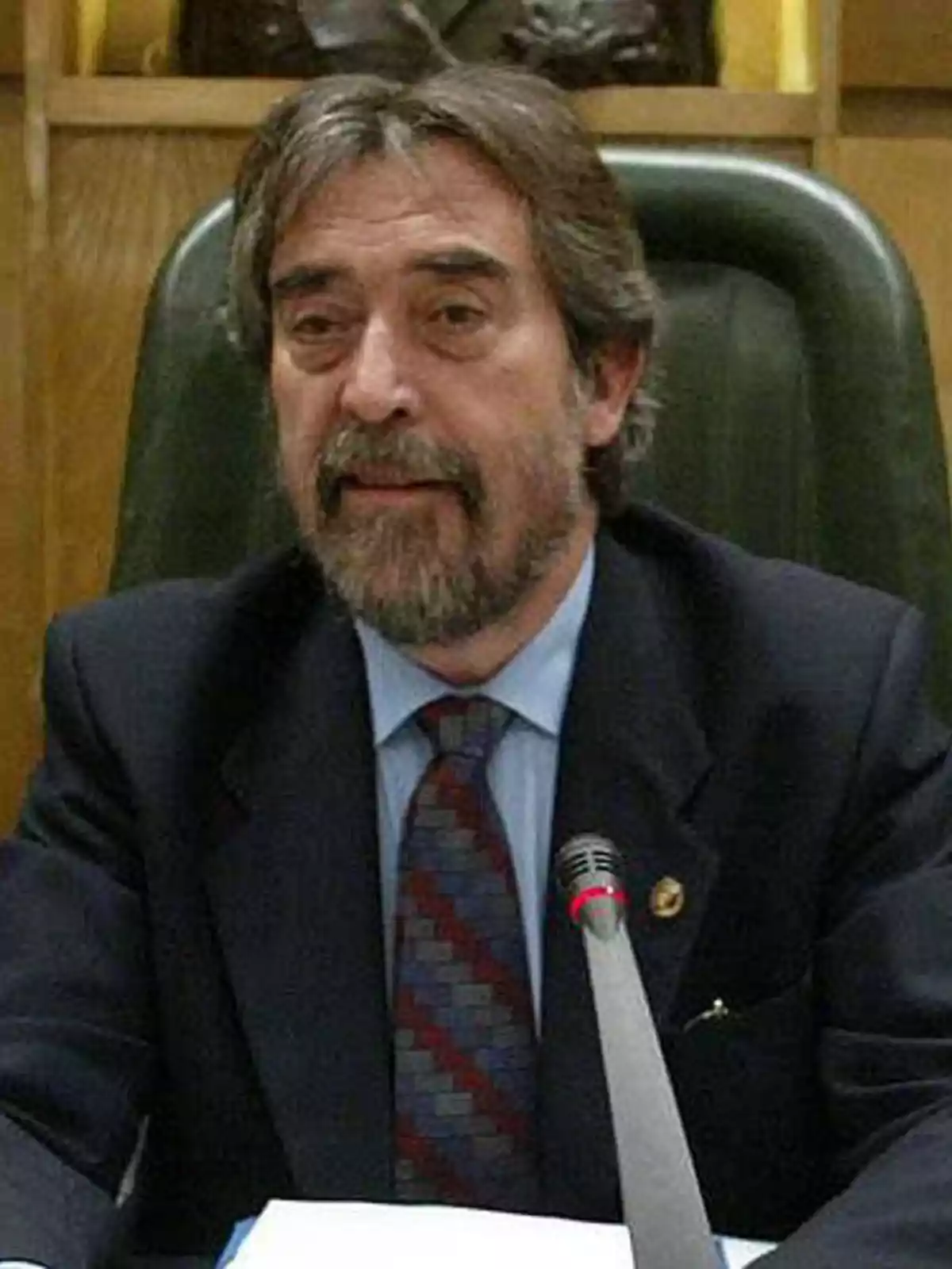 Hombre de mediana edad con barba y cabello canoso sentado en una silla grande, usando traje y corbata, hablando frente a un micrófono en un entorno formal Hombre de mediana edad con barba y cabello canoso sentado en una silla grande, usando traje y corbata, hablando frente a un micrófono en un entorno formal