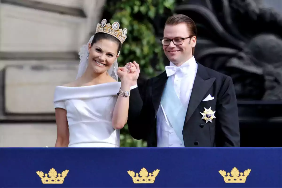 Pareja vestida de gala sonríe y saluda desde un balcón decorado con coronas doradas Pareja vestida de gala sonríe y saluda desde un balcón decorado con coronas doradas