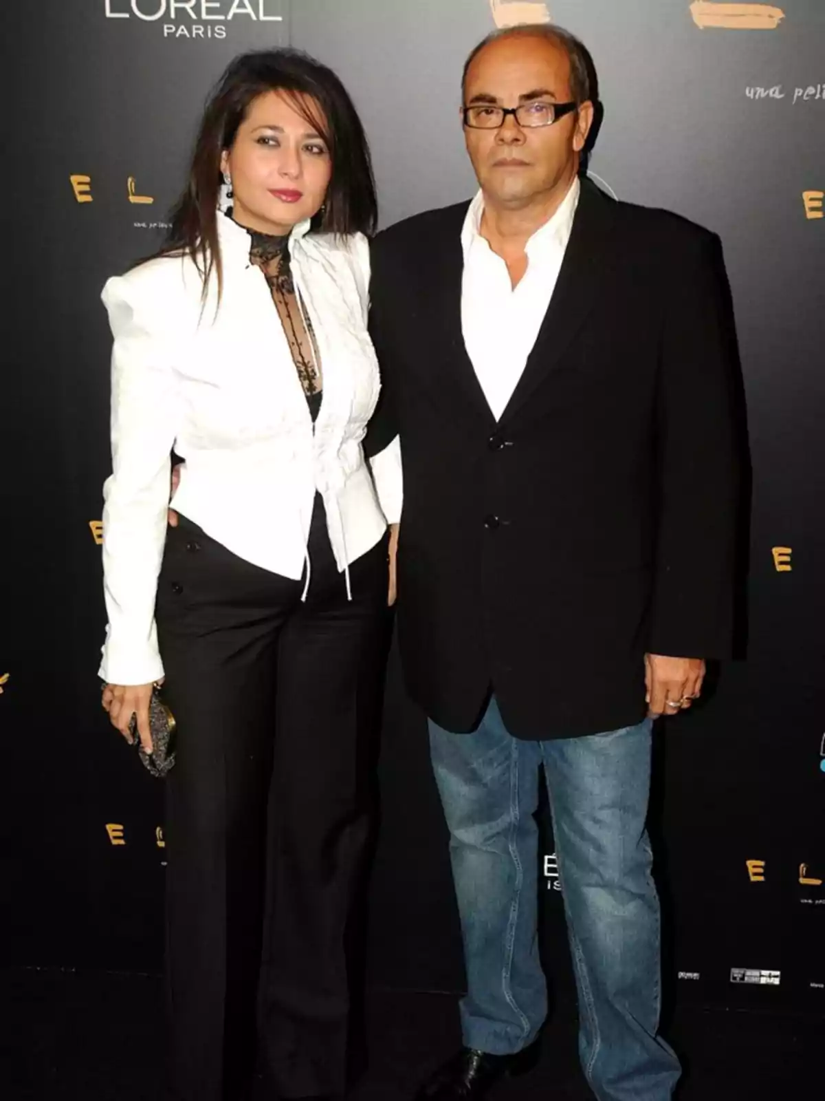 Una mujer y un hombre posan juntos en una alfombra roja con fondo negro y logotipos de marcas, ambos vestidos de manera elegante.