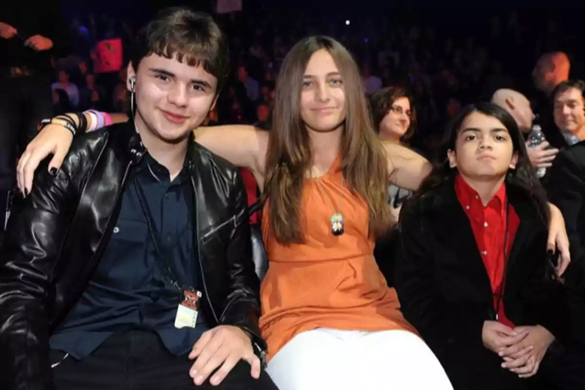 Tres jóvenes sentados juntos en un evento, la persona del centro lleva un vestido naranja y abraza a los otros dos, todos miran hacia la cámara con expresiones relajadas
