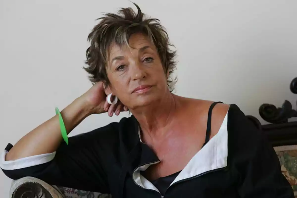 Mujer de cabello corto y canoso sentada en un sillón con expresión pensativa, usando aretes blancos y una pulsera verde Mujer de cabello corto y canoso sentada en un sillón con expresión pensativa, usando aretes blancos y una pulsera verde