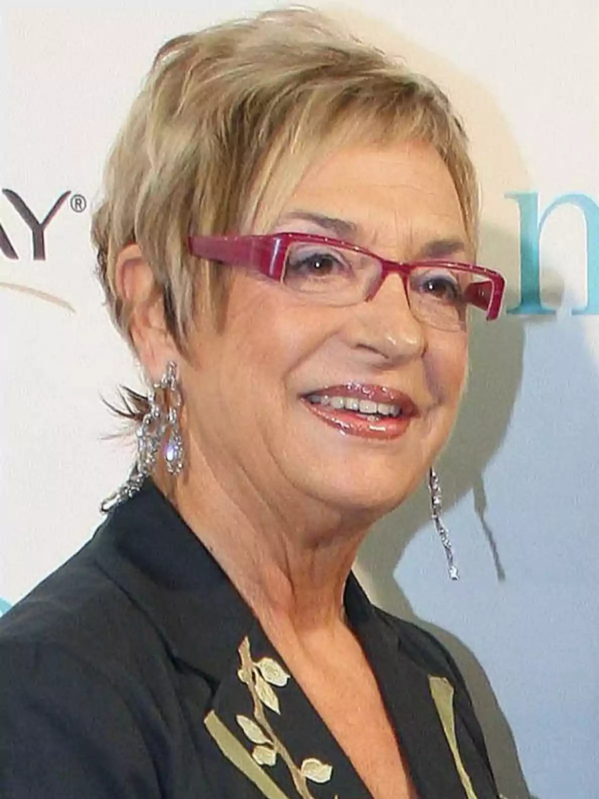 Mujer mayor con gafas rojas, cabello corto rubio y pendientes largos, sonriendo y usando un saco oscuro con detalles claros Mujer mayor con gafas rojas, cabello corto rubio y pendientes largos, sonriendo y usando un saco oscuro con detalles claros