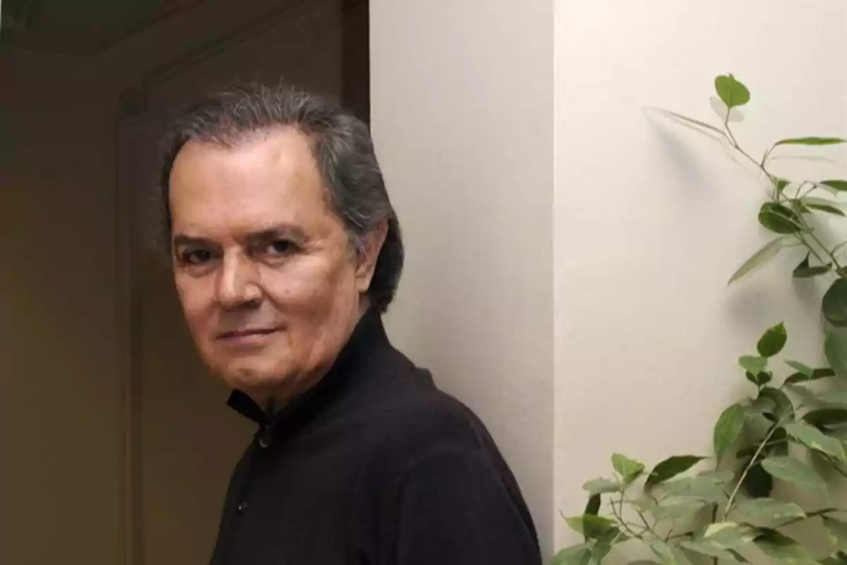 Hombre de cabello canoso y expresión seria, vestido con camisa oscura, de pie junto a una pared blanca y una planta verde. Hombre de cabello canoso y expresión seria, vestido con camisa oscura, de pie junto a una pared blanca y una planta verde.