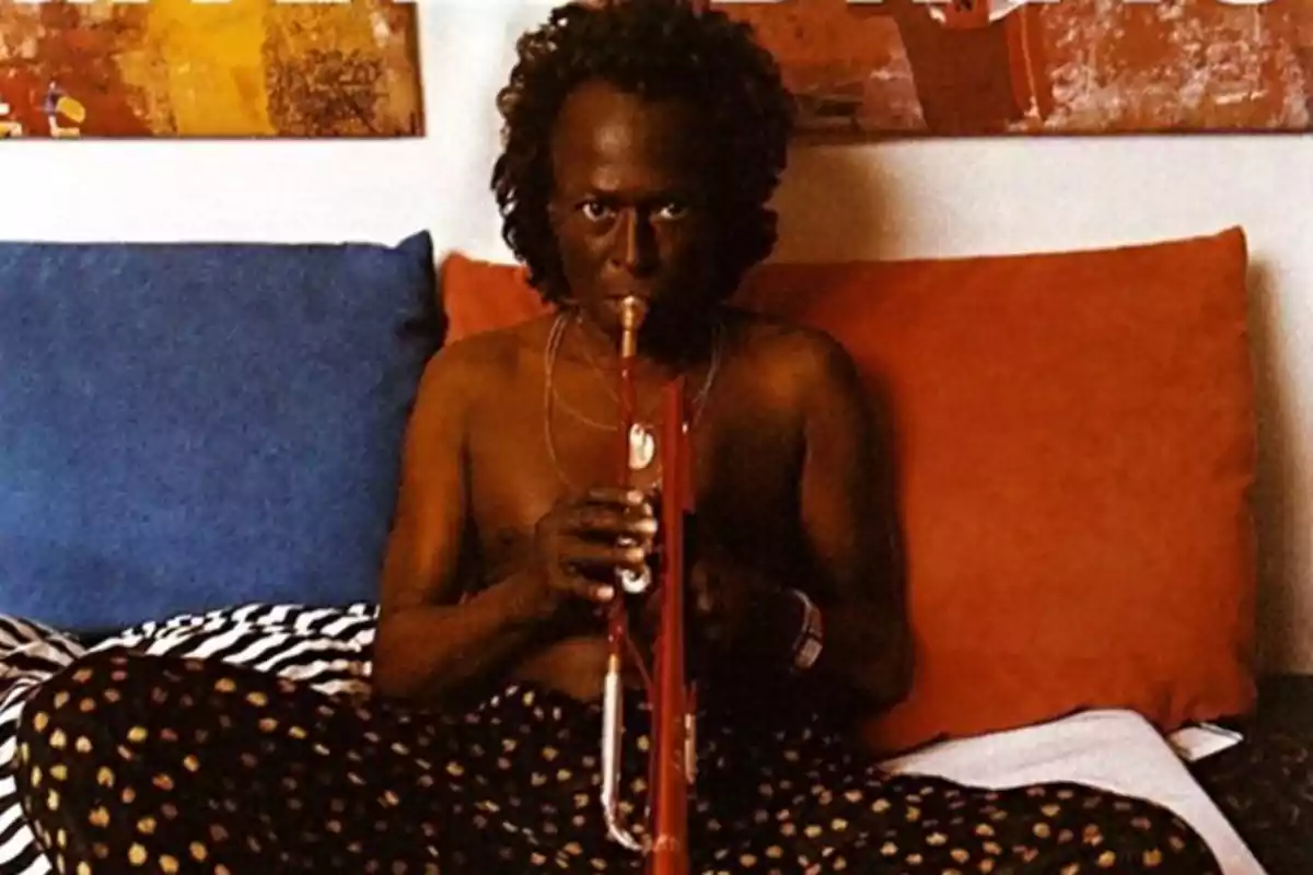 Hombre sentado en un sofá tocando una trompeta roja sin camisa con cojines de colores y una pintura abstracta al fondo