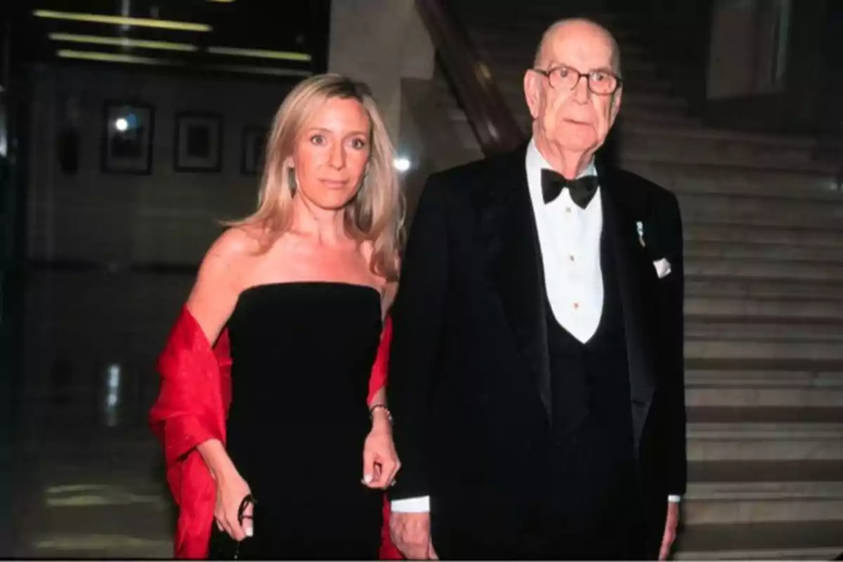 Una mujer con vestido negro y chal rojo junto a un hombre mayor con esmoquin y pajarita posan en una escalera elegante Una mujer con vestido negro y chal rojo junto a un hombre mayor con esmoquin y pajarita posan en una escalera elegante