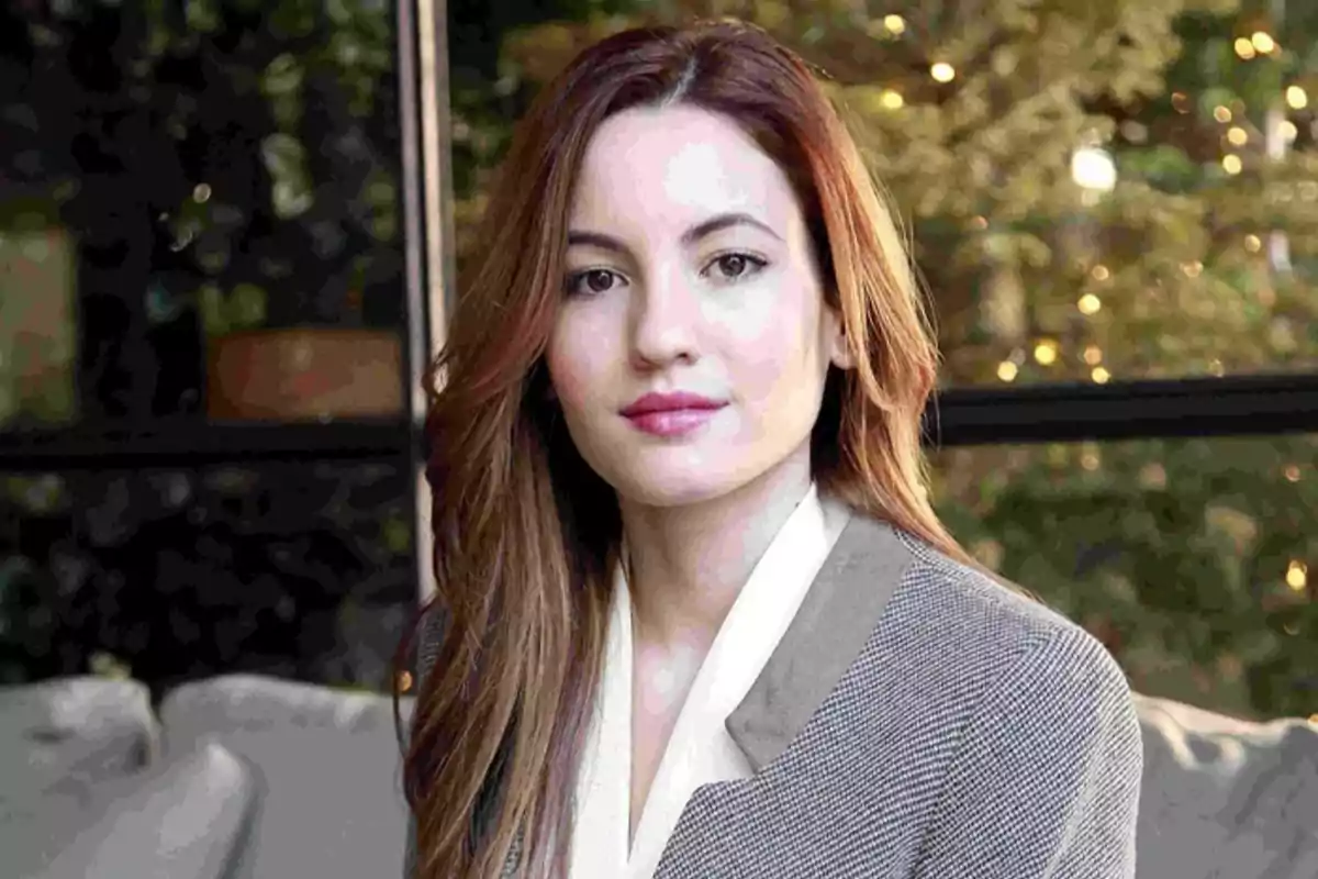 Mujer joven de cabello castaño claro y largo con saco gris y blusa blanca sentada en un ambiente interior con fondo de luces y plantas Mujer joven de cabello castaño claro y largo con saco gris y blusa blanca sentada en un ambiente interior con fondo de luces y plantas