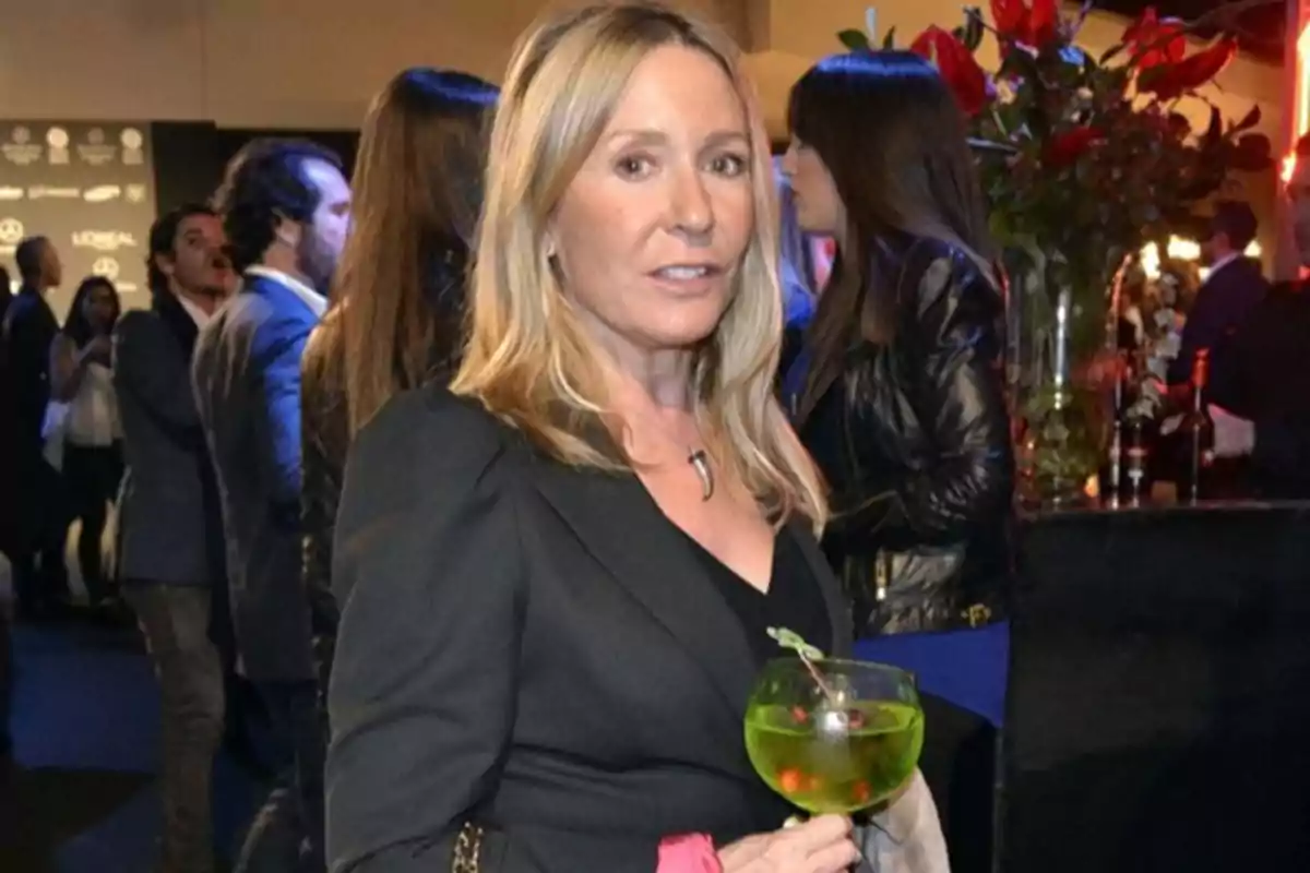 Mujer rubia con vestido negro sosteniendo una copa de cóctel verde en un evento social con varias personas al fondo Mujer rubia con vestido negro sosteniendo una copa de cóctel verde en un evento social con varias personas al fondo