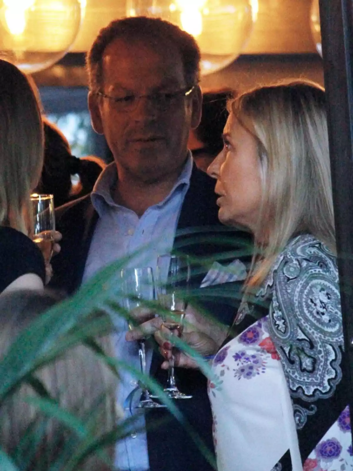 Dos personas conversan en una reunión social mientras sostienen copas de vino, rodeadas de otras personas y luces cálidas en el fondo Dos personas conversan en una reunión social mientras sostienen copas de vino, rodeadas de otras personas y luces cálidas en el fondo