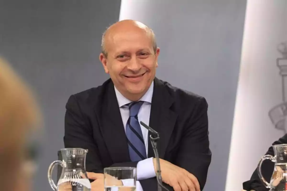 Hombre calvo con traje oscuro y corbata azul sonriendo mientras está sentado en una mesa con micrófono y jarras de agua