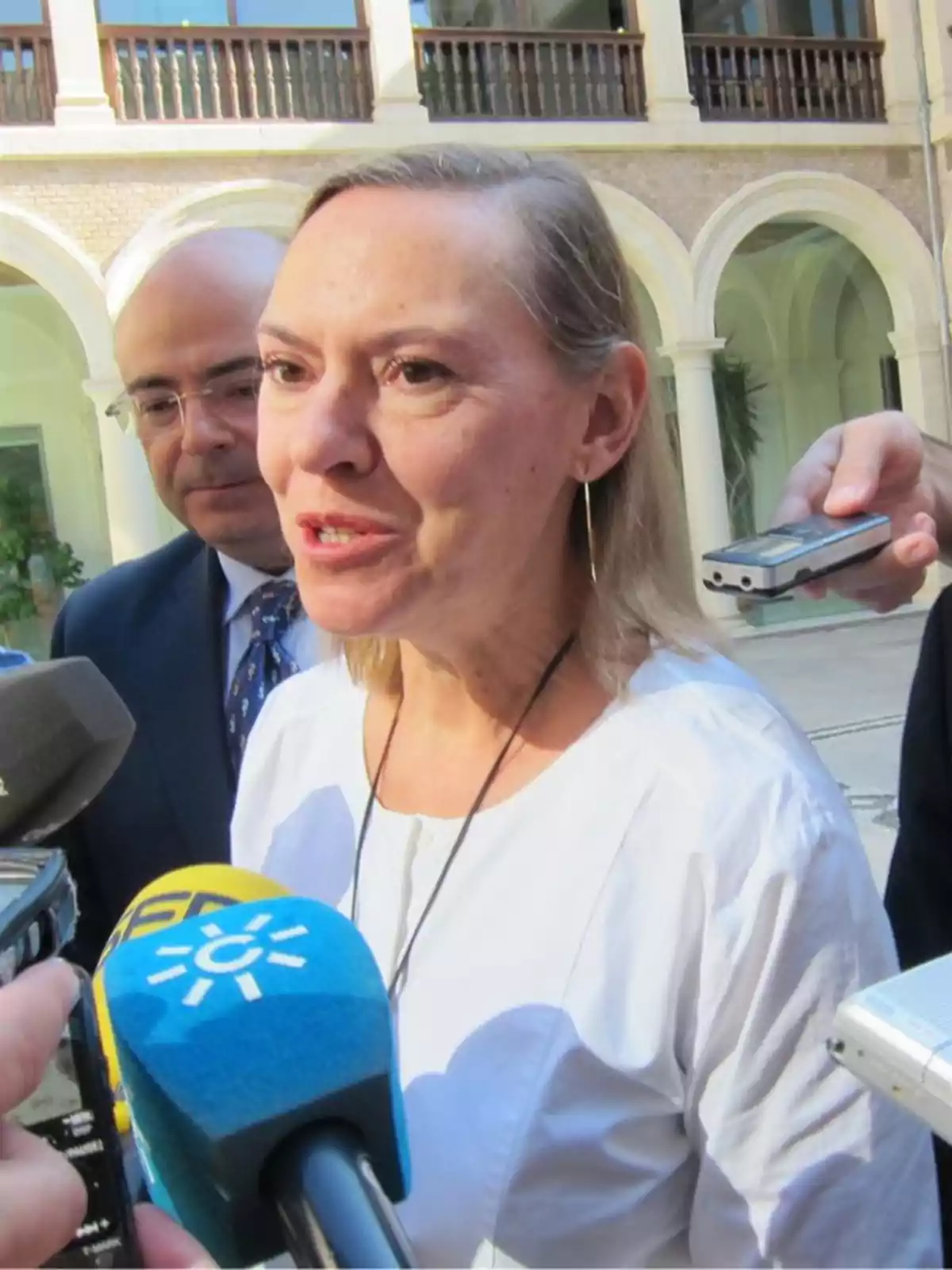 Mujer de cabello rubio y corto vestida con blusa blanca dando declaraciones a la prensa rodeada de micrófonos y grabadoras en un patio con arcos Mujer de cabello rubio y corto vestida con blusa blanca dando declaraciones a la prensa rodeada de micrófonos y grabadoras en un patio con arcos