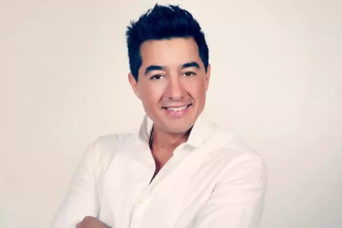 Hombre de cabello oscuro con camisa blanca sonriendo frente a un fondo claro Hombre de cabello oscuro con camisa blanca sonriendo frente a un fondo claro