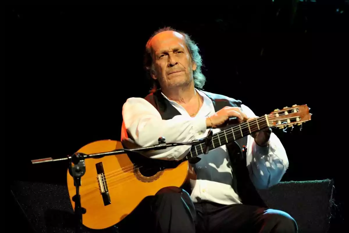 Hombre mayor sentado en el escenario tocando una guitarra española bajo una luz cálida