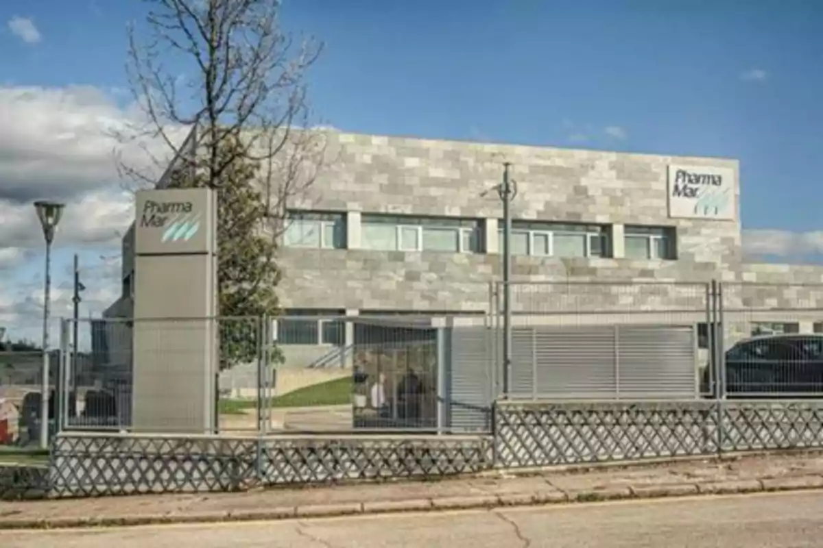 Edificio moderno de dos plantas con fachada de piedra gris y letreros de PharmaMar rodeado por una reja metálica y un árbol al frente bajo un cielo parcialmente nublado Edificio moderno de dos plantas con fachada de piedra gris y letreros de PharmaMar rodeado por una reja metálica y un árbol al frente bajo un cielo parcialmente nublado