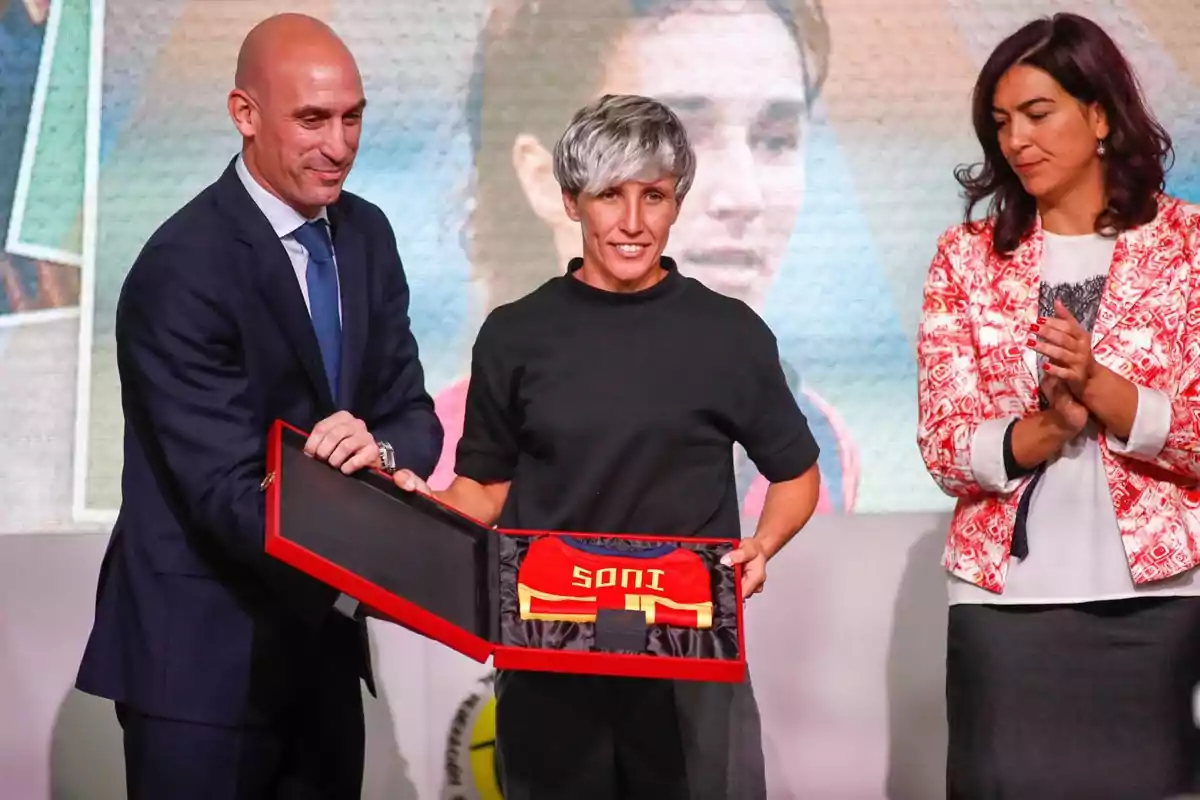 Tres personas en un evento formal, una de ellas sostiene una caja con una camiseta roja y amarilla que tiene el nombre Soni, mientras otra persona aplaude Tres personas en un evento formal, una de ellas sostiene una caja con una camiseta roja y amarilla que tiene el nombre Soni, mientras otra persona aplaude