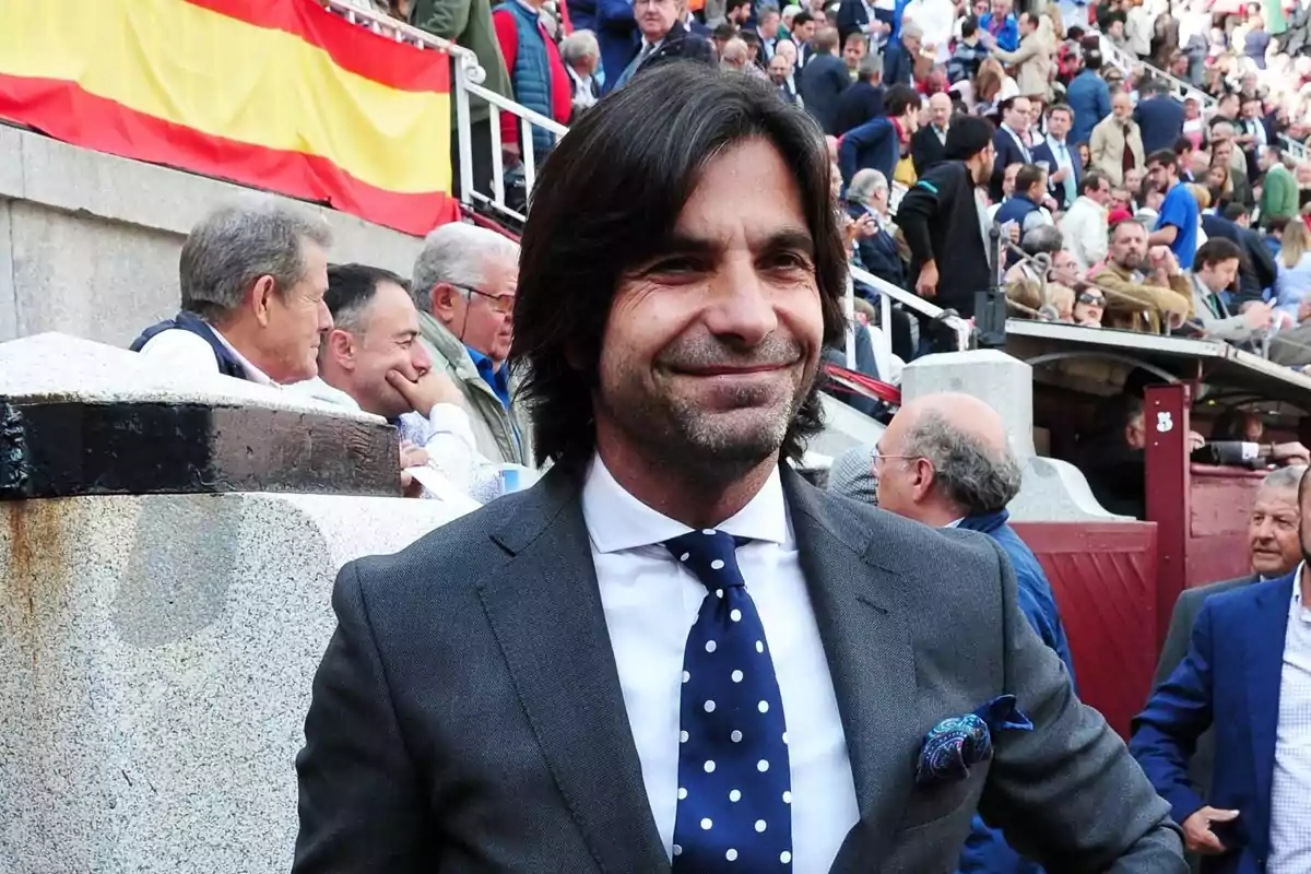 Hombre de cabello oscuro y largo con traje gris y corbata de lunares sonriendo en una plaza de toros llena de gente