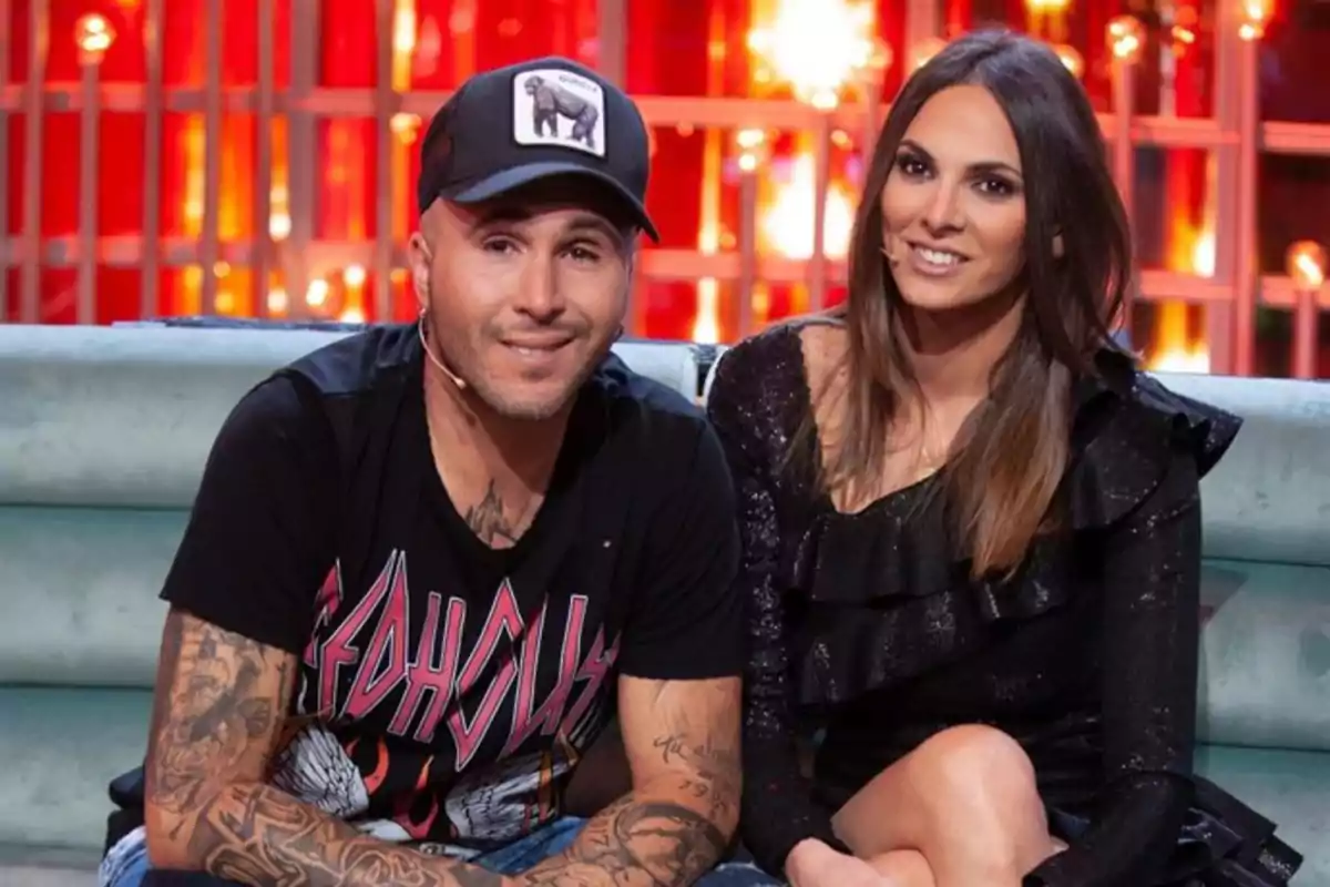 Un hombre con gorra y camiseta negra con tatuajes en los brazos está sentado junto a una mujer de cabello largo y liso vestida de negro en un sofá, ambos sonríen y están en un set de televisión con luces rojas de fondo Un hombre con gorra y camiseta negra con tatuajes en los brazos está sentado junto a una mujer de cabello largo y liso vestida de negro en un sofá, ambos sonríen y están en un set de televisión con luces rojas de fondo