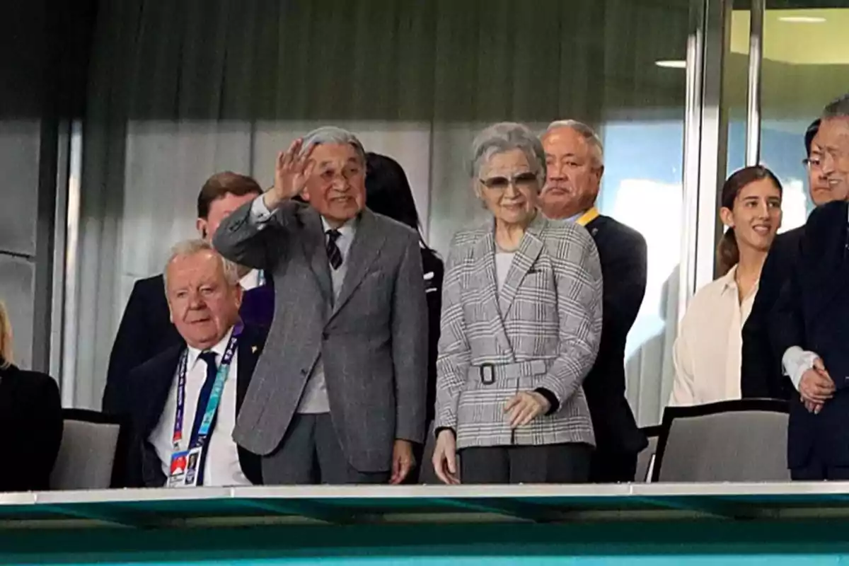 Un hombre mayor saluda con la mano mientras está de pie junto a una mujer mayor con gafas y otras personas en un palco de un estadio Un hombre mayor saluda con la mano mientras está de pie junto a una mujer mayor con gafas y otras personas en un palco de un estadio