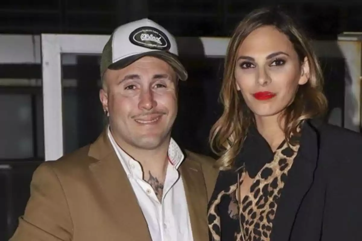 Un hombre con chaqueta marrón y gorra blanca posa junto a una mujer de cabello suelto y labios rojos que lleva un abrigo negro y blusa de estampado animal