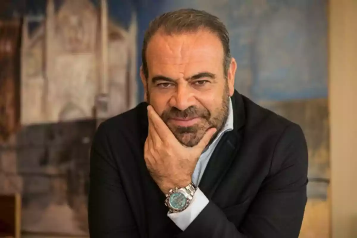 Hombre de barba y cabello corto con saco oscuro y reloj plateado apoyando la barbilla en su mano frente a un fondo desenfocado Hombre de barba y cabello corto con saco oscuro y reloj plateado apoyando la barbilla en su mano frente a un fondo desenfocado