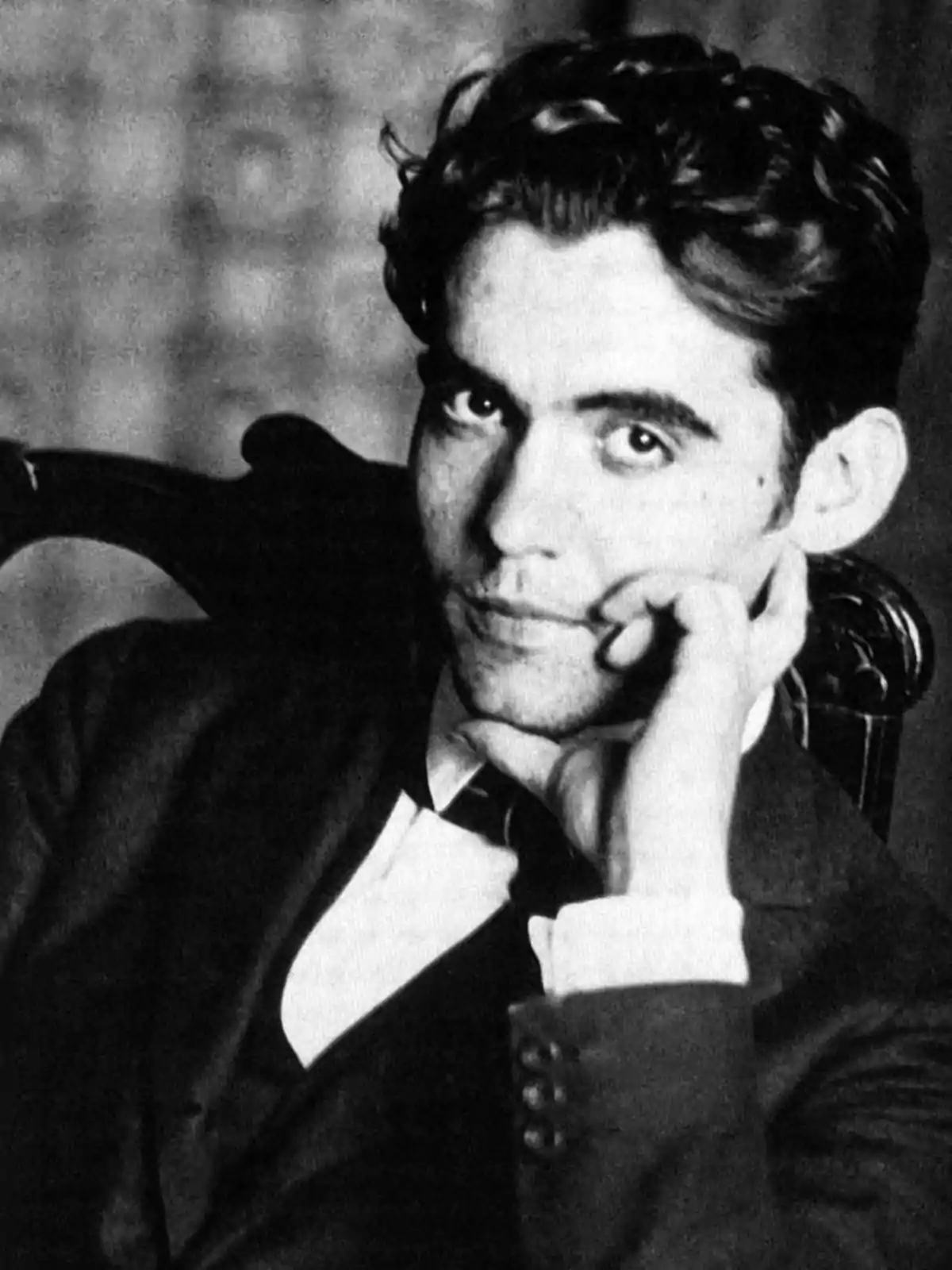 Retrato en blanco y negro de un hombre joven con cabello oscuro y ondulado, vestido con traje y corbata, apoyando la cabeza en su mano y mirando a la cámara con expresión pensativa Retrato en blanco y negro de un hombre joven con cabello oscuro y ondulado, vestido con traje y corbata, apoyando la cabeza en su mano y mirando a la cámara con expresión pensativa