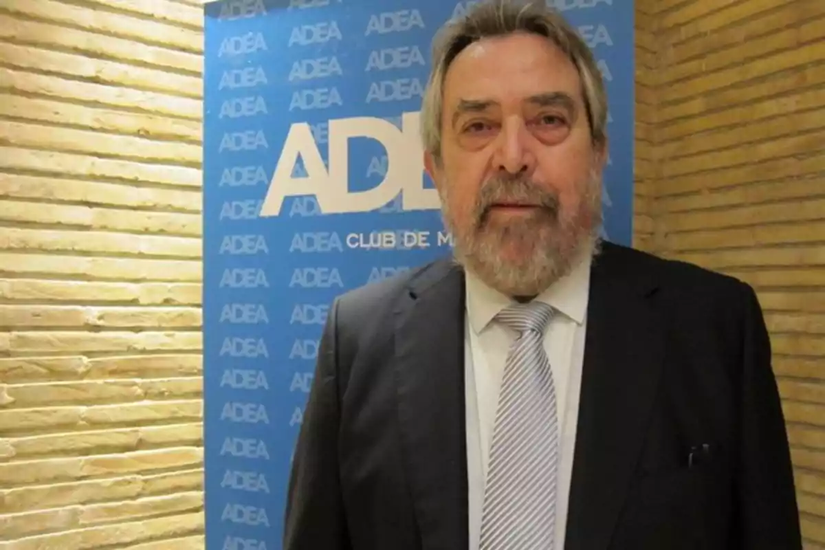 Hombre de cabello y barba canosos con traje y corbata posando frente a un fondo azul con el logo de ADEA Hombre de cabello y barba canosos con traje y corbata posando frente a un fondo azul con el logo de ADEA