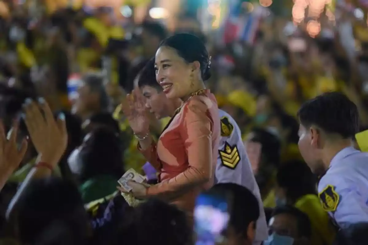 Mujer vestida con ropa tradicional sonríe y saluda entre una multitud de personas, acompañada por oficiales con uniforme blanco Mujer vestida con ropa tradicional sonríe y saluda entre una multitud de personas, acompañada por oficiales con uniforme blanco