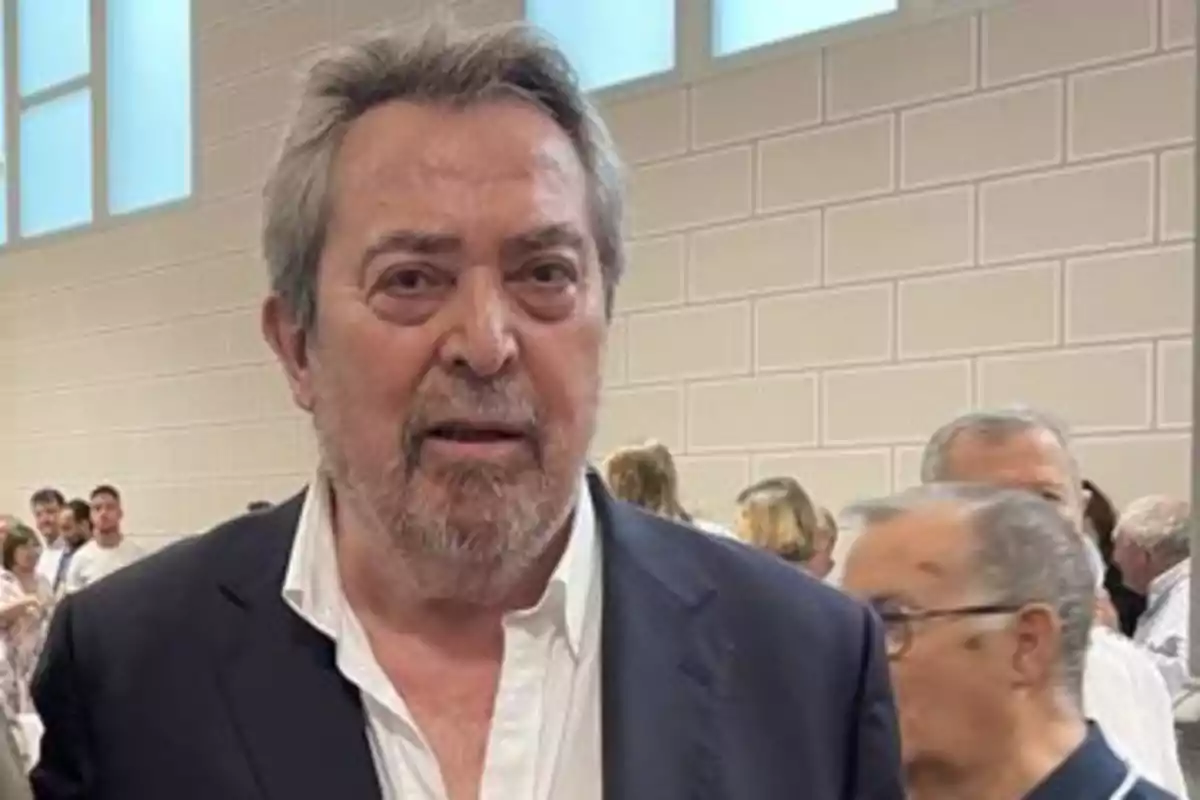 Hombre de cabello canoso y barba con saco oscuro y camisa blanca en un evento social en un recinto cerrado con varias personas al fondo Hombre de cabello canoso y barba con saco oscuro y camisa blanca en un evento social en un recinto cerrado con varias personas al fondo