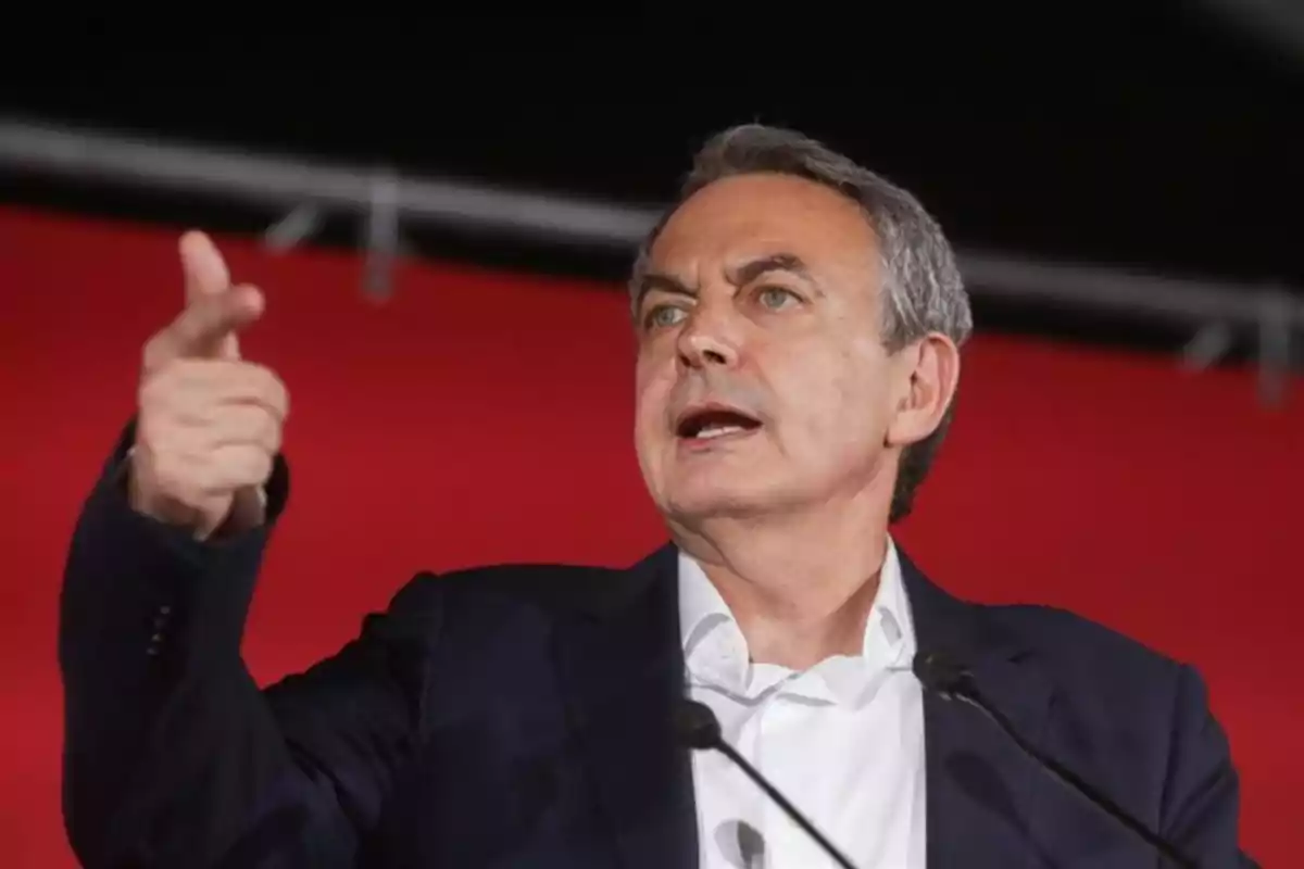 Hombre de cabello canoso con traje oscuro hablando en un podio y gesticulando con la mano frente a un fondo rojo