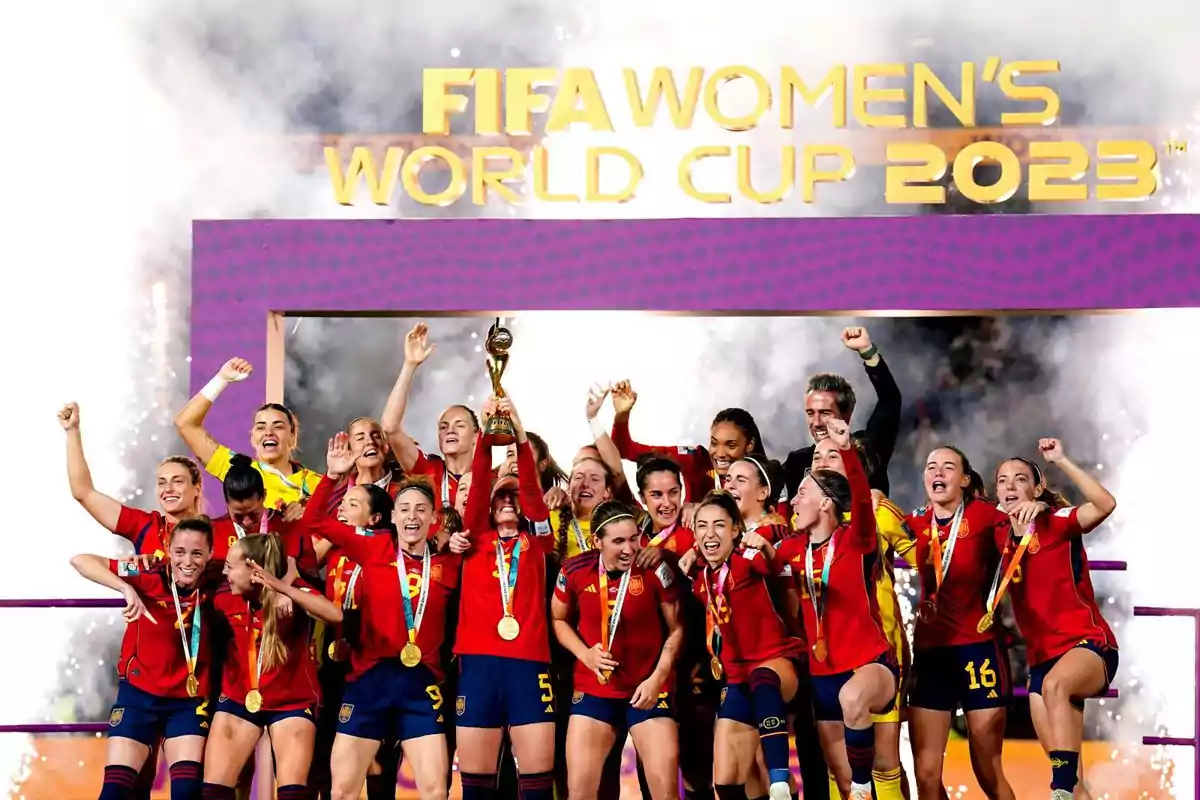 Jugadoras de la selección española de fútbol femenino celebran con medallas y el trofeo tras ganar la Copa Mundial Femenina 2023 bajo un letrero dorado y fuegos artificiales de fondo