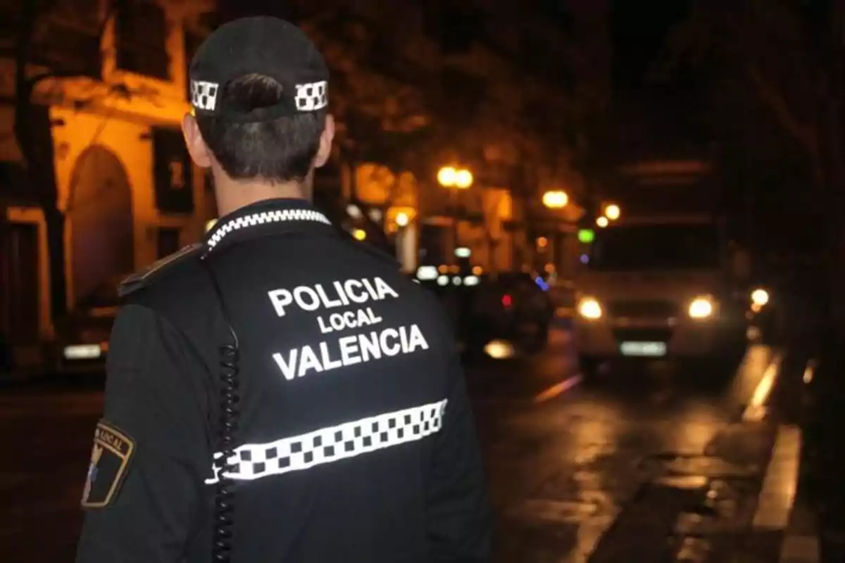 Agente de la policía local de Valencia de espaldas vigilando una calle por la noche con luces de vehículos encendidas al fondo Agente de la policía local de Valencia de espaldas vigilando una calle por la noche con luces de vehículos encendidas al fondo