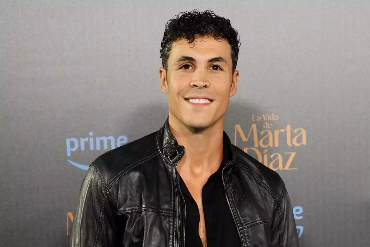 Hombre joven de cabello rizado y chaqueta de cuero negro sonriendo frente a un fondo con logotipos de Prime Video y La Vida de Marta Díaz Hombre joven de cabello rizado y chaqueta de cuero negro sonriendo frente a un fondo con logotipos de Prime Video y La Vida de Marta Díaz