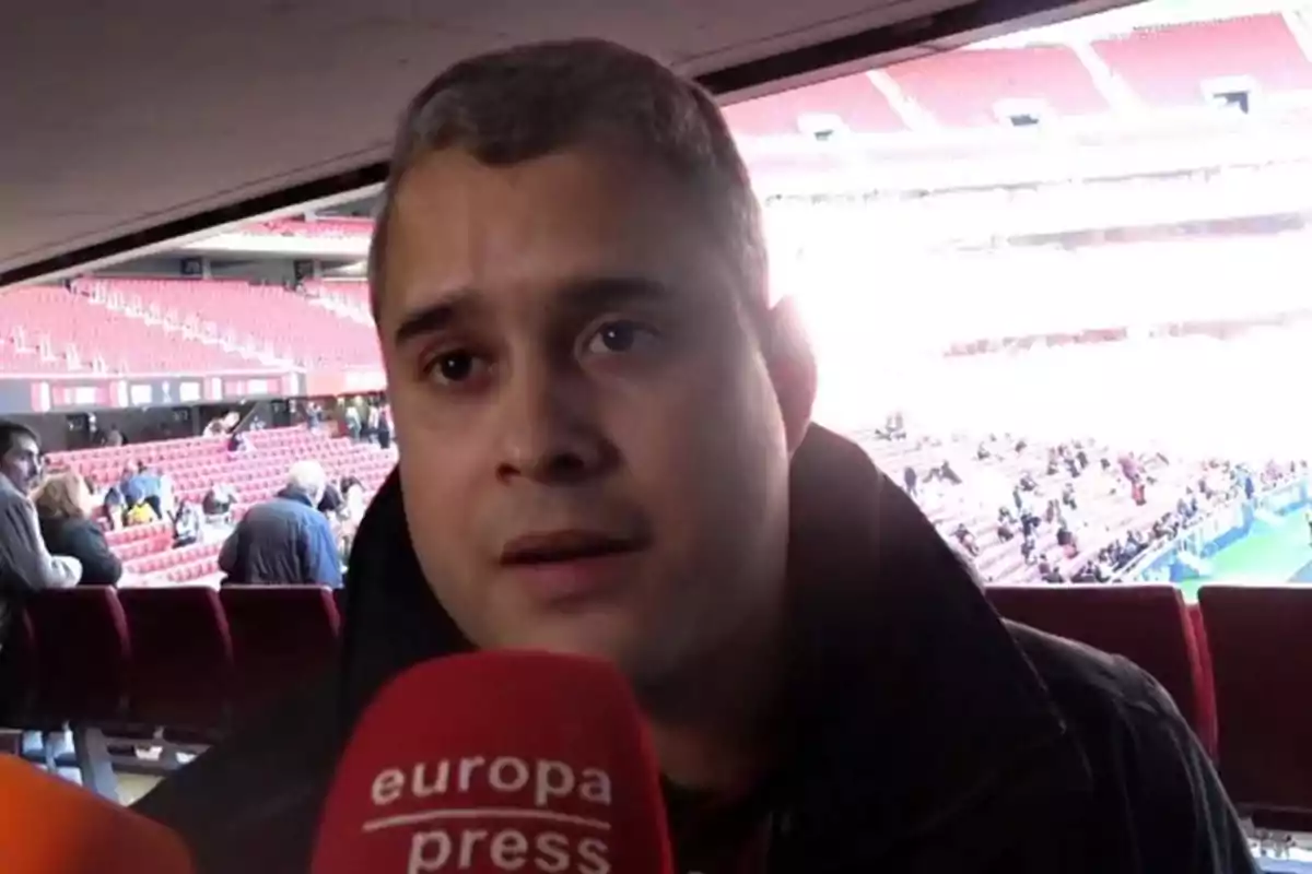 Un hombre es entrevistado en un estadio de fútbol con gradas rojas y micrófonos frente a él Un hombre es entrevistado en un estadio de fútbol con gradas rojas y micrófonos frente a él