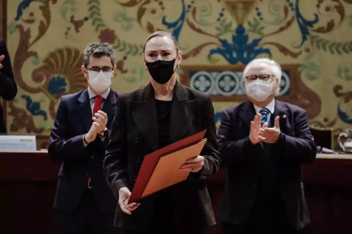 Mujer con mascarilla negra sostiene un diploma mientras dos hombres con mascarilla blanca la aplauden en un evento formal con fondo decorativo. Mujer con mascarilla negra sostiene un diploma mientras dos hombres con mascarilla blanca la aplauden en un evento formal con fondo decorativo.