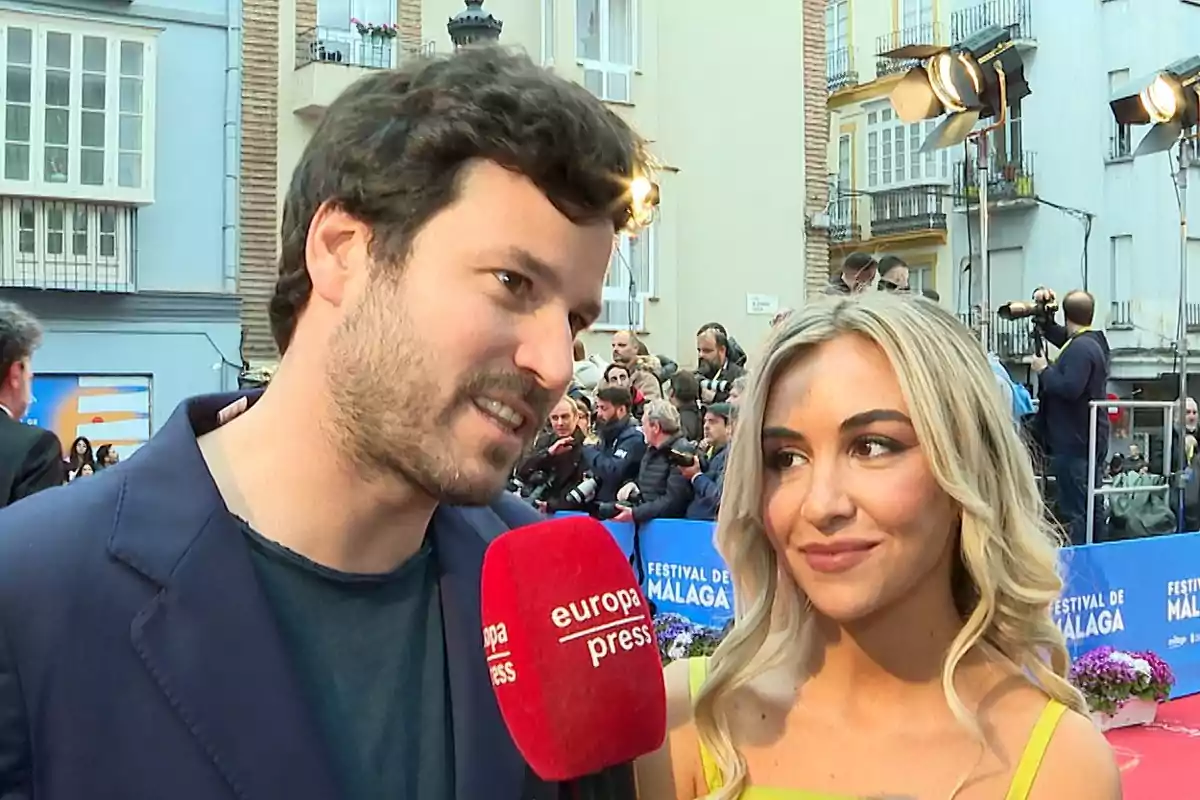 Un hombre y una mujer son entrevistados en una alfombra roja durante un evento al aire libre con público y fotógrafos al fondo Un hombre y una mujer son entrevistados en una alfombra roja durante un evento al aire libre con público y fotógrafos al fondo