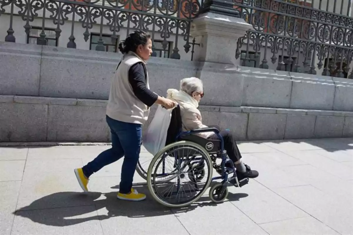 Una mujer empuja a una persona mayor en silla de ruedas por una acera junto a una reja de hierro y un muro de piedra Una mujer empuja a una persona mayor en silla de ruedas por una acera junto a una reja de hierro y un muro de piedra