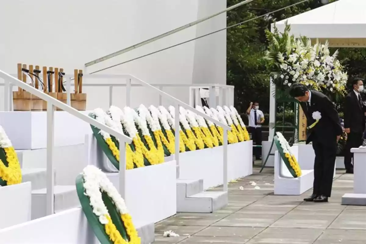 Hombre vestido de traje negro inclina la cabeza frente a una ofrenda floral en una ceremonia con coronas de flores blancas y amarillas Hombre vestido de traje negro inclina la cabeza frente a una ofrenda floral en una ceremonia con coronas de flores blancas y amarillas