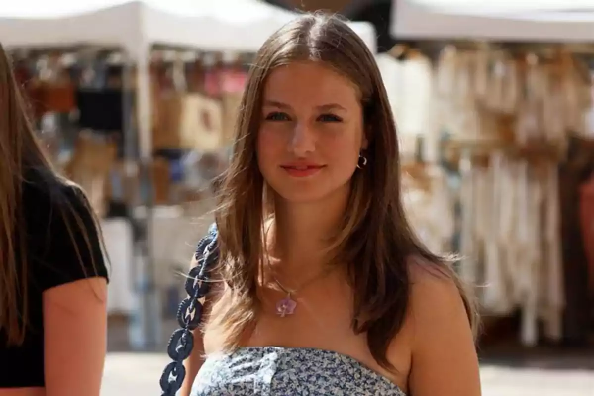 Young woman with straight light brown hair, wearing a blue strappy dress and a necklace, smiling outdoors at a craft market. Young woman with straight light brown hair, wearing a blue strappy dress and a necklace, smiling outdoors at a craft market.